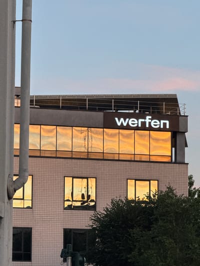 The image shows the facade of a building in Sesto San Giovanni, Italy, at dusk. The building is several stories high and features large windows that reflect the warm, golden light of the setting sun, creating abstract patterns. A prominent sign with the illuminated white text "werfen" is affixed to the top section of the building. The building's exterior is clad in light-colored square tiles. To the left, a large, light-colored pipe runs vertically along the building. In the foreground and to the right, dense green foliage from trees partially obscures the lower floors of the building. The sky above is a soft blue with a hint of pink.