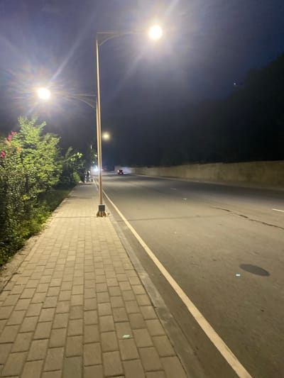 The image depicts a nighttime scene on a paved road with a sidewalk. The road has a white line marking the lane. The sidewalk is made of light-colored rectangular pavers. There are streetlights illuminating the area, creating bright glare. Green foliage and some pink flowers are visible along the edge of the sidewalk. In the distance on the road, there appear to be a few vehicles, possibly motorcycles or scooters, with their headlights on. The sky is dark, indicating nighttime, and there are some subtle lights visible in the upper portion of the frame. The overall atmosphere is that of a quiet, urban or suburban street at night, illuminated by artificial lighting.
