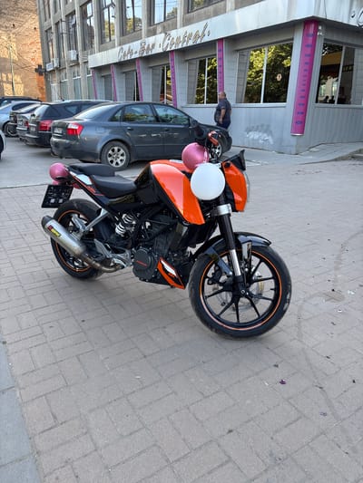 The image shows an orange and black KTM motorcycle parked on a paved street in Hayredin, Bulgaria. The motorcycle has two balloons attached to it: one pink and one white. Behind the motorcycle are parked cars and a building with multiple windows and a sign that reads "Cafe-Bar Central". A person is partially visible standing behind the cars, looking towards the motorcycle. Debris is scattered on the pavement near the front wheel of the motorcycle.