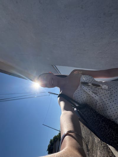 A person is taking a selfie in the city of Bulgarovo, Bulgaria. The person is wearing a white polka dot dress and holding a black bag. They are standing outside on a sunny day, with a bright sunburst in the background. The person is smiling and their face is partially obscured by the sun. The environment includes a concrete structure overhead, power lines, and a clear blue sky. The image is taken from a low angle, looking upwards.