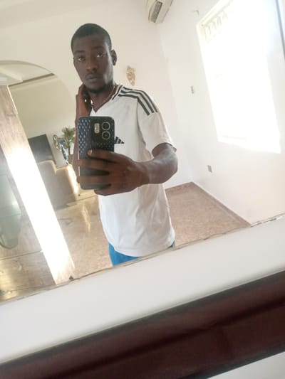 A man is taking a selfie in a brightly lit room. He is wearing a white t-shirt with black stripes on the sleeves and blue shorts. He is holding a smartphone in his right hand, with the camera facing himself. The background shows a mirror reflecting the room, which includes a light-colored wall, a window with bright sunlight streaming through, and a light brown tiled floor. In the reflection, a portion of a couch and a fan are visible. The image appears to have been taken in a residential setting in Hotoro North, Nigeria.