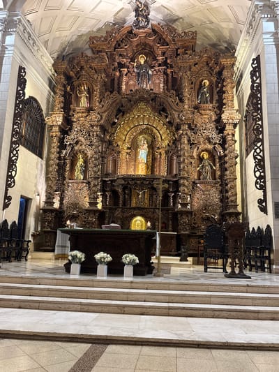This image depicts the opulent interior of the San Isidro Church in Lima, Peru. The central focus is the ornate, gilded altarpiece, a towering structure intricately carved from dark wood, adorned with sculptures of religious figures, including a prominent depiction of the Virgin Mary holding the child Jesus. The altarpiece is illuminated by spotlights, casting a warm glow on its detailed craftsmanship. In front of the altarpiece is a dark wooden altar, draped with white fabric and adorned with three white floral arrangements in square white vases. A single lit candle sits on the altar. To the left of the altar, a dark wooden lectern is visible. The floor in front of the altar is a polished marble with a patterned tile inlay. Several rows of tiered marble steps lead up to the altar area. Dark wooden chairs with high backs are placed on either side of the sanctuary. The church's ceiling is high and features a patterned coffered design. The overall impression is one of solemnity and grandeur, highlighting the rich religious and artistic heritage of the location.