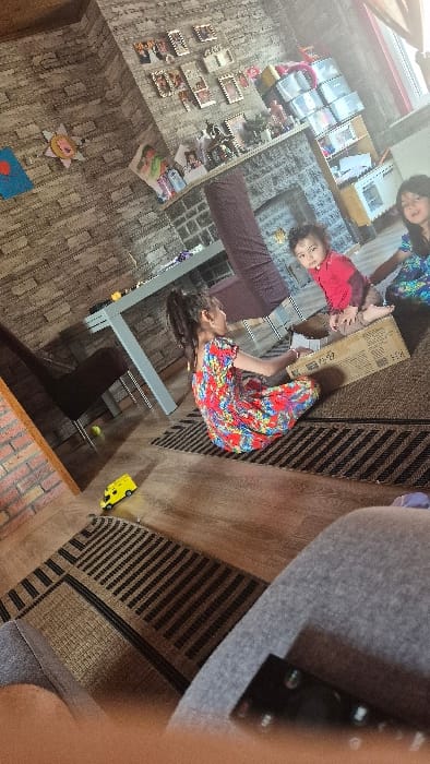This image captures an indoor residential scene, specified as being in Melle, Belgium. Three children are present in a room featuring light-toned faux stone wall paneling on the right, contrasted with a brick wall section on the left. The floor is comprised of wooden planks partially covered by two rectangular dark brown and beige striped rugs.
In the center-midground, a young girl, wearing a multi-colored floral dress with her dark hair in a ponytail, is seated on one rug, facing away from the viewer. She is positioned next to a large cardboard box. Inside this box, a young child or toddler, wearing a red long-sleeved shirt and light trousers, is seated and looking directly towards the camera. To the right and slightly behind the box, another young girl, wearing a blue and white patterned dress, is partially visible, also looking towards the camera.
In the background, a light-colored rectangular table with a dark chair and a purple fabric-covered chair are visible. A fireplace mantel below a collection of framed pictures and decorative items is against the faux stone wall. A window on the far right allows natural light into the room. On the left wall, a yellow sun-shaped decoration and a blue art piece are affixed. A yellow toy vehicle is on the floor to the left, and a small green ball is near the dark chair. The bottom right foreground shows a portion of a grey fabric surface, likely a sofa or lap, with a remote control partially visible.