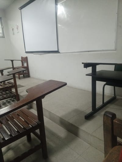 This image captures an empty classroom or lecture hall in Santa Cruz de la Sierra, Bolivia. The scene features rows of desks and chairs, primarily made of dark wood, arranged for students. Each wooden desk has an integrated chair with a slatted back and seat, and a flat writing surface extending to the right. In the foreground, one such wooden desk and chair is prominently visible, while others recede into the background on the left. On the right side of the room, a single black metal desk with an attached black chair stands out, possibly designated for an instructor or a different type of student seating.

The front wall of the classroom is painted a light color, likely white or off-white. A large retractable projector screen is pulled down, covering a significant portion of the wall, with a separate whiteboard visible to its right. Reflections on the whiteboard suggest an overhead light source or natural light. To the left of the projector screen, a window with horizontal panes allows natural light into the room.

Two square signs are affixed to the wall near the window. One clearly depicts a "No Smoking" symbol, featuring a red circle with a diagonal line over a cigarette. The other sign, to its left, is less distinct but also appears to be a prohibitory sign with "NO" and some symbols within a red circle, possibly indicating "No Food" or "No Drinks."

The floor is covered with light-colored, square tiles, possibly grey or beige. There's a subtle elevation or step running across the floor in the midground of the room. No people are present, and no activity is taking place, giving the room a quiet, unoccupied atmosphere. The lighting is moderate, suggesting daytime.