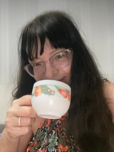 A woman with long, dark hair and glasses holds a small, white teacup with a floral design. She is wearing a ring on her left hand and a patterned top. The background is a plain, light-colored wall. The image appears to be a selfie taken indoors in Novi Petrivtsi, Ukraine.