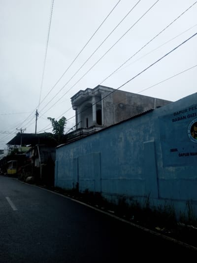 The image depicts a static street scene under an overcast sky, suggesting a cloudy day, early morning, or late afternoon. No people are visible.

In the foreground, a dark asphalt road with a faded white line on its left edge runs diagonally upwards from the bottom left corner. To the right of the road, a long, tall, light blue wall dominates the midground. This wall appears textured and slightly weathered, with faint rectangular indentations. Over the wall, a corrugated metal roof is partially visible.

Above the blue wall, a multi-story building with a distinctive architectural style can be seen. It features a prominent upper section with classical columns and ornate molding along its roofline, suggesting a formal or older design. Further back and to the left of this building, other, less distinct structures are visible, along with green foliage, possibly banana leaves. Utility poles with numerous power lines crisscross the upper portion of the frame against the grey sky, converging towards the left horizon.

Visible text on the blue wall, written in Indonesian, includes: "DAPUR PEL. BADAN GIZI" (likely "Dapur Pelayanan Badan Gizi" - Nutrition Service Agency Kitchen or similar), "REPUBLIK INDONESIA" (Republic of Indonesia), and partially visible "YAYA DAPUR MAN." (possibly related to a foundation or independent kitchen). Adjacent to the text is the emblem of the Republic of Indonesia, the Garuda Pancasila. These textual and emblematic cues firmly indicate the location is Indonesia, not Babaliya Mal., India, as previously suggested.