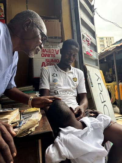 This media file shows an outdoor scene in Ikoyi, Nigeria. Two men are the subjects of the image. One man, with grey hair and glasses, is wearing a light blue medical scrub top and looking upwards and to the right. He has a goatee and appears to be speaking or observing something out of frame. The other two men are younger and appear to be interacting with each other, possibly in a market setting. One man is wearing a white polo shirt with the text "LAGOS" and "FirstBank" visible, and the other is wearing a white garment. They are lying down or leaning on a wooden surface.
In the background, there are shelves filled with what appear to be books or documents. There are also signs, including one that reads "Pay" and another with "Google Play" and an "ORANGE" brand visible on a box. A corrugated metal structure is also prominent, suggesting an open-air market or stall. The weather appears overcast. The overall impression is of a busy, possibly informal, commercial area. The text suggests a business or service-oriented environment. The interaction between the two younger men, with one appearing to be resting, and the man in the medical scrub top looking upwards, could imply a pause in their day or a conversation.