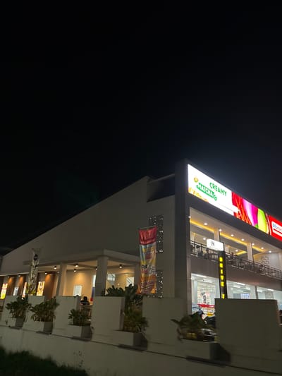 An exterior nighttime low-angle shot captures a modern, multi-story building with bright signage. The building, likely a commercial establishment, is illuminated, creating a warm glow from within. A prominent, illuminated sign displays "CREAMY MATCHA &" in large green lettering, with colorful abstract graphics to its right. To the far right, a red sign with white lettering reads "Alfamart."
A balcony level is visible on the second floor, where several people are seated at tables. A sign with the letters "IN" is also illuminated on this level. Further down, the ground floor appears to be a retail space, possibly a convenience store, with shelves and products visible through large glass windows. A motorcycle is parked outside.
Along the front of the building, planters with lush green foliage are positioned in front of white rectangular planters. Decorative banners hang vertically on the left and right sides of the building, featuring text and graphics that are partially visible. The sky above is dark, suggesting it is nighttime.
This building is located in Tangerang, Indonesia, as indicated by the presence of an "Alfamart" store, a widely recognized convenience store chain in Indonesia.
