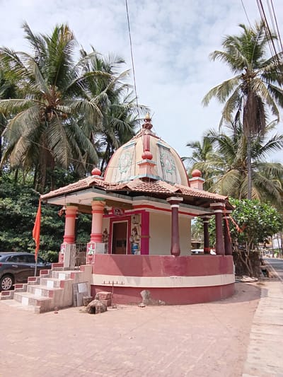The image depicts a small, colorful Hindu temple in Chopdem, India, situated amidst palm trees. The temple has a dome-shaped roof adorned with decorative patterns and a red flag flying nearby. The structure is painted in shades of red, white, and pink, with pillars and a railing surrounding the main shrine. A paved pathway leads to the temple entrance. A car is visible parked on the side of the road adjacent to the temple steps. The sky is overcast, suggesting a cloudy day.