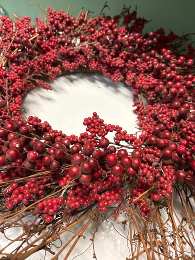 This media file is a close-up, vertically oriented, outdoor photograph of a wreath made of red berries and brown twigs. The wreath is circular and is arranged on a surface that appears to be a white wall or backdrop, with a subtle shadow cast by the wreath. The lighting suggests an indoor setting, possibly a home or a craft store. The wreath is densely packed with clusters of small, red berries and interspersed with thin, dried brown twigs and vines. Some of the berries are glossy, while others have a matte finish, indicating they might be artificial. The texture of the berries is varied, with some appearing smooth and round, and others slightly elongated or clustered together. The overall impression is one of a rustic, decorative item, likely intended for holiday or seasonal display. There is no visible text in the image. The focus is entirely on the detailed texture and arrangement of the wreath's components. The image does not depict any people, animals, or specific environmental conditions like weather. The implied location of Bradenton, United States, provides a general geographical context but does not offer specific visual cues within the image itself.