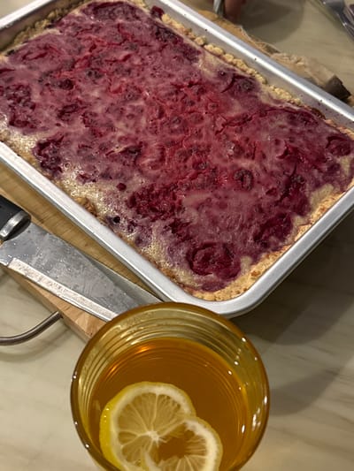 An overhead shot shows a rectangular berry tart in a metal baking pan, with a glass of amber liquid containing lemon slices in the foreground. The tart appears to have a golden-brown crust and is topped with a thick layer of red berries and a slightly bubbly, translucent glaze. The baking pan is positioned on a wooden cutting board, next to which is a chef's knife with a black handle. The setting appears to be a kitchen counter or table. The image does not provide information about its location in San Phra Net Subdistrict, Thailand.