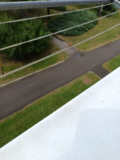 This media file is an outdoor, medium-angle shot looking downwards. The foreground is dominated by a white, slightly textured surface, possibly a railing or balcony edge. Beyond this, a grey metal pipe runs horizontally across the top of the frame.
Below the pipe and railing, the scene unfolds. Four thin, parallel wires stretch across the image, creating a layered effect over the landscape. The landscape consists of vibrant green grass, a dark asphalt path or road, and several dense green bushes. The path appears to have some wet patches, suggesting recent rain or dew. In the background, there are hints of trees and more distant greenery.
The overall impression is of an urban or suburban setting, likely a park or residential area with landscaping and paved pathways. The lighting suggests it is daytime, and the weather appears to be overcast or possibly just after rain, given the wet surfaces. There are no people or animals visible in the scene. The focus is on the arrangement of man-made elements like the railing, pipe, wires, and path against the natural backdrop of grass and bushes. The composition is simple, with a clear distinction between the immediate foreground and the background elements. No visible text is present in the image. The location context of Košice, Slovakia, places this scene within a European urban environment.