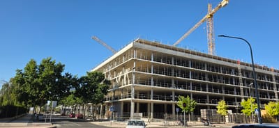 An outdoor, eye-level, wide shot captures a large, multi-story building under construction in Huesca, Spain. Two yellow construction cranes are visible against a clear blue sky. The building, composed of concrete and scaffolding, has multiple open floors with visible support columns and railings.
In the foreground, a paved street curves to the left, with a pedestrian sidewalk to the right of the street. Several green trees line the sidewalk, with one particularly large tree dominating the left side of the frame. Red and silver cars are parked or driving on the street. A white car is visible on the sidewalk near the lower right corner of the image.
Further down the sidewalk, a street lamp with a curved arm stands tall. Signs on poles, including a blue pedestrian crossing sign and a green sign with "ATENCION STOP," are present. Stacked materials, likely construction supplies, are visible on the right side of the sidewalk. A parked scooter is also present on the sidewalk. The scene suggests a sunny day in an urban or suburban environment undergoing development.