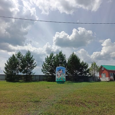 The image depicts a rural scene in Orlovka, Russia, under a partly cloudy sky. In the foreground, a grassy field stretches out, with some patches of dry grass indicating the time of year is likely summer or early autumn. A prominent object in the middle of the field is a large blue cylindrical structure adorned with a whimsical painting of cows, sunflowers, and a picket fence, suggesting it might be a water tank or a decorative element for a farm or village.
Behind the field, a line of tall pine trees forms a backdrop. Beyond the trees, a metal fence is visible. To the right of the frame, a red brick house with a green roof stands, featuring windows and a small white square attic window. The house appears to be a residential dwelling. Power lines crisscross the sky, a common sight in rural areas.
The sky is dramatic, with bright shafts of sunlight breaking through fluffy white cumulus clouds, casting shadows and creating a dynamic play of light and dark. The overall mood is serene and bucolic, evoking a sense of peaceful country life. The time of day appears to be late morning or early afternoon, given the angle of the sunlight. There is no visible text in the image.