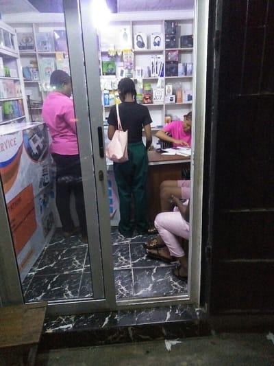 An interior view of a retail store, likely an electronics or mobile phone accessory shop, in Cotonou, Benin, is captured through a silver-framed glass entrance. The time appears to be evening or night due to the dark exterior.

Inside, the store features white display shelves along the back wall, stocked with various electronic products including headphones, speakers, cables, and other packaged accessories. The floor is tiled with a black and white marble-patterned design.

Four individuals are present. On the far left, a female is standing with her back to the camera, wearing a pink long-sleeved shirt and dark trousers. To her right, another female stands with her back to the camera, wearing a black t-shirt, dark green baggy trousers, and carrying a light pink patterned tote bag over her right shoulder.

On the right side of the store, a wooden counter is visible. A third female is seated at this counter, facing partially away from the camera, wearing a bright pink t-shirt with striped sleeves. She is engaged in writing or reviewing papers laid out on the counter. Below the counter, a fourth individual is seated, partially obscured, with only their legs and feet clearly visible. This person is wearing light pink trousers and dark sandals, and appears to be interacting with a mobile device held in their hands.

A promotional stand or counter with "SERVICE" and "TATION" visible on a poster is located on the left inside the entrance. Outside the entrance on the lower left, a small wooden stool is positioned on a dark, rough ground surface, where some white debris is present.