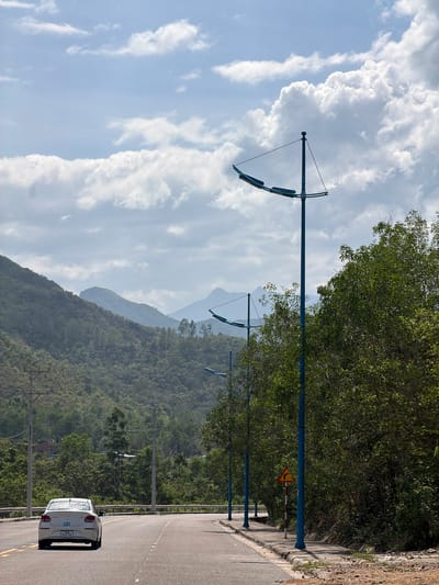 A white car is driving on a paved road with yellow lane markings. The road curves to the left, and on the right side of the road, there is a sidewalk with blue lampposts and a yellow warning sign indicating a sharp curve to the right. A dense green forest covers the hills in the background, and the sky is filled with white and gray clouds. The scene appears to be on the outskirts of Vĩnh Lương, Vietnam, possibly along a scenic route.