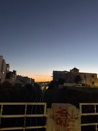 The image captures a twilight scene in Cascais, Portugal, likely overlooking a railway line. In the foreground, a white railing, partly obscured by graffiti, borders the viewpoint. Beyond the railing, multiple railway tracks recede into the distance, flanked by dark, sloped embankments. Along the crest of the embankments, buildings with various architectural styles are visible, some featuring distinct rooflines and windows. In the mid-ground, construction cranes are present, suggesting ongoing development or infrastructure work. The sky transitions from a deep blue at the top to a vibrant orange near the horizon, indicating the sun has recently set. A single, distant star or planet is visible in the upper right quadrant of the sky. The overall atmosphere is calm and urban, with hints of industrial activity due to the railway and cranes.