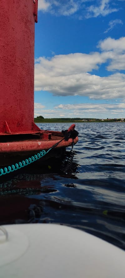 The image shows a close-up view of a red buoy floating on dark blue water. A coiled turquoise rope is attached to the buoy and extends into the water. In the background, there is a clear blue sky with white clouds, a line of green trees, and several small houses in the distance. The location is identified as Щиглицы, Russia. The scene appears to be a calm day on a body of water, possibly a lake or a river, with a buoy marking a navigation channel.