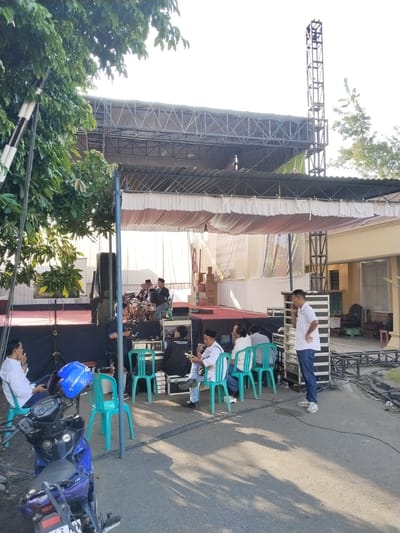 This outdoor scene captures the preparation or a rehearsal for an event, likely a musical performance, in Sragen, Indonesia, under bright daylight.
In the center, a large temporary stage is set up, featuring a metal truss structure supporting a roof and a white fabric backdrop. On the stage, a drummer and other musicians are visible, surrounded by musical equipment and speakers. Stacked cardboard boxes, some labeled "TOSHIBA," are also on the stage.
To the right of the stage, a tall metal tower stands, likely for lighting or sound. Below it, stacks of sound equipment and mixers are arranged. A man in a white polo shirt and jeans stands next to this equipment, looking towards the stage, possibly managing the sound.
Several people are seated on green plastic chairs near the stage. Some are observing the stage, while others appear to be conversing. One man on a chair wears a white shirt and a black peci/kufi. A blue motorcycle is parked in the foreground, with a blue helmet displaying "Evolution" resting on its handlebar. Part of its license plate, "63 AH," is visible.
The ground is paved asphalt with cables running across it. Trees with lush green foliage frame the left side of the image and appear in the background to the right. A building with yellow walls and metal grates on its windows is visible behind the stage on the right. The strong sunlight casts clear shadows on the ground, indicating either morning or late afternoon. A partially visible banner behind the stage has text, including phrases like "MENYAMBUT KEPUTUSAN YUDISIUM..." and "...DEKAN SEWUN".