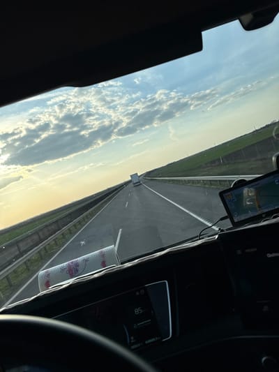 The image depicts a highway scene from the perspective of a vehicle's driver, likely a truck, traveling at an indicated speed of 85 km/h. The setting is consistent with the M43 motorway in the vicinity of Makó, Hungary, characterized by flat, open agricultural fields bordering the road.
The sky exhibits scattered cumulus clouds under a bright, low sun on the left, suggesting late afternoon or early morning. The highway ahead is a multi-lane asphalt road with clear white markings, extending into the distance. Metal guardrails separate the highway from the green fields on both sides. A white semi-trailer truck is visible in the distance, proceeding in the same direction.
Inside the vehicle, the dashboard is visible, featuring a digital instrument cluster displaying "85" (presumably km/h) and an odometer reading of "408689.3 km". A portable navigation device is mounted on the right side of the dashboard, showing a map interface and also indicating a speed of 85 km/h. A roll of white paper towels with a purple/pink grape motif rests on the dashboard in front of the instrument panel. The top rim of a steering wheel is visible at the bottom left. No individuals are visible in the frame.