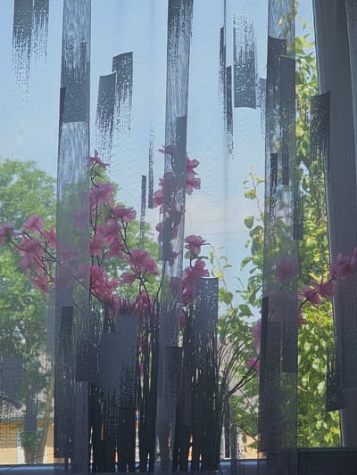 The image shows a close-up of a sheer curtain with a patterned design, obscuring a view of outdoor foliage and the sky. The curtain has vertical dark grey rectangular shapes with speckled patterns, interspersed with horizontal lines throughout its fabric. Through the semi-transparent curtain, blurred pink flowers on slender stems are visible in the mid-ground, alongside lush green leaves and branches of trees. The background appears to be a bright sky. The overall scene suggests an indoor perspective looking out through a window covered by this decorative curtain. The location is identified as Dūkštas, Lithuania.