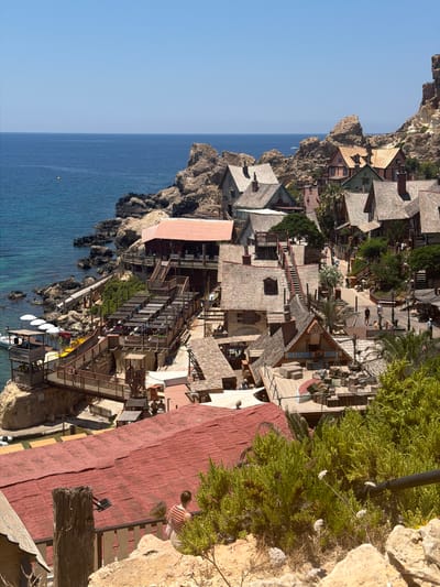 The image depicts Popeye Village, a small amusement park and tourist attraction located in the North of Malta, near the town of Mellieħa. The village is situated on a rocky coastline overlooking the Mediterranean Sea. The scene is characterized by a collection of colorful, wooden buildings with thatched roofs, resembling a cartoonish fishing village.
In the foreground, there are several wooden structures, including a large building with a red roof and a wooden balcony. A person wearing a striped shirt is visible on this balcony. Various wooden walkways and stairs connect different parts of the village.
Further into the scene, more buildings are visible, many with different colored facades and roofs. Some of these buildings are set against the natural backdrop of rocky cliffs. There are several people walking around the village, suggesting it is open to visitors.
The sea is a prominent feature, with its clear blue water lapping against the rocky shore. A small boat is docked near the water's edge, and some white umbrellas are set up on a platform. The overall atmosphere is bright and sunny, with a clear blue sky. The architecture and setting strongly suggest this is a film set or themed attraction, likely based on the Popeye comics.