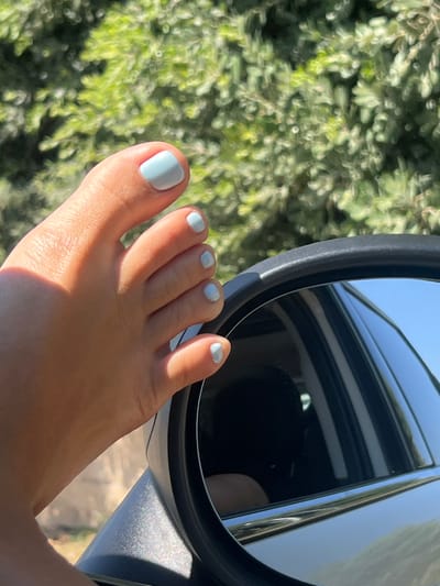 A person's bare foot, with pale blue painted toenails, rests on the side-view mirror of a car. The foot, positioned from a close-up perspective, shows its toes and the sole of the foot illuminated by sunlight, creating a shadow on the side. The background consists of out-of-focus green foliage and a clear blue sky reflected in the car's mirror, which also shows a glimpse of the car's interior. The image does not provide specific details to determine the location within the city of Fasano, Italy.