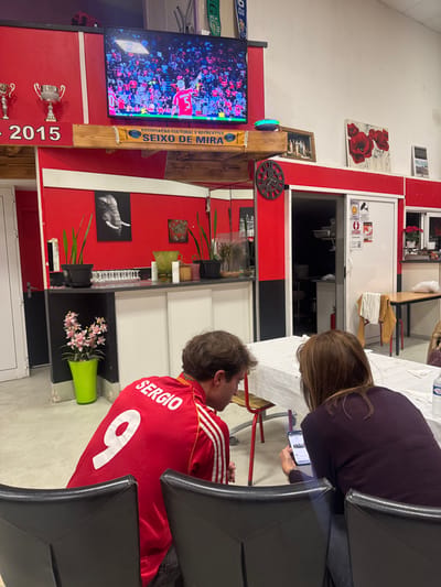 The image depicts an indoor scene within the venue of the "ASSOCIAÇÃO CULTURAL E RECREATIVA SEIXO DE MIRA," located in Décines-Charpieu, France.
The environment features red and white walls with a light-colored floor. A large flat-screen television is mounted high on a red wall, displaying a live soccer match featuring a player in a red jersey with number 5. Below the television, a blue and yellow banner with the association's name is displayed on a wooden shelf. To the left of the television, two silver-colored trophies are visible, with "2015" text below them.
A bar area is present with a white countertop. On the left section of the bar, multiple clear glasses are arranged, flanked by two potted snake plants. Above this, a framed black and white image of an elephant's head hangs on the red wall. On the floor to the left of the bar, a bright green pot holds pink artificial flowers. On the right section of the bar, another potted plant and a small glass aquarium are visible. A dark, ornate clock with exposed gears is mounted on the red wall to the right of the bar.
To the right, a white sliding door, partially open, reveals a dark interior, likely a kitchen. The door has multiple regulatory signs affixed, including a CCTV symbol and "STOP COVID-19" information. Above this door, a painting featuring red poppies on a white background, along with another framed picture, adorns the upper wall. A small shelf near the kitchen entrance holds a red poinsettia plant and another item.
In the foreground, two individuals are seated at a white-tablecloth-covered table, viewed from behind. The person on the left is a male with medium brown hair, wearing a red soccer jersey with "SERGIO" and the number "9" printed in white on the back, along with white Adidas stripes on the sleeves. He is looking down, towards the smartphone held by the second person. The person on the right is a female with brown, shoulder-length hair, wearing a dark purple top. She is actively looking at and interacting with a smartphone held in her hands. Both are seated on dark-colored chairs.