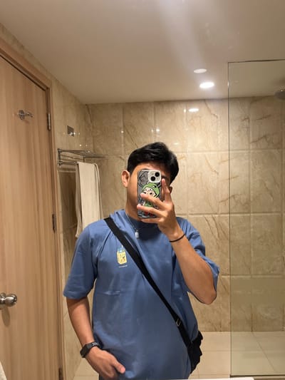 A young man stands in a bathroom, holding up a smartphone to take a mirror selfie. He is wearing a blue t-shirt with a cartoon graphic and a black crossbody bag. His right arm is bent, resting on his stomach, and his left hand is in his pocket. A black fitness tracker is visible on his left wrist. The bathroom features marble-tiled walls, a wooden door with a silver handle, and a towel rack with a beige towel. The lighting suggests an indoor setting, possibly a hotel room.