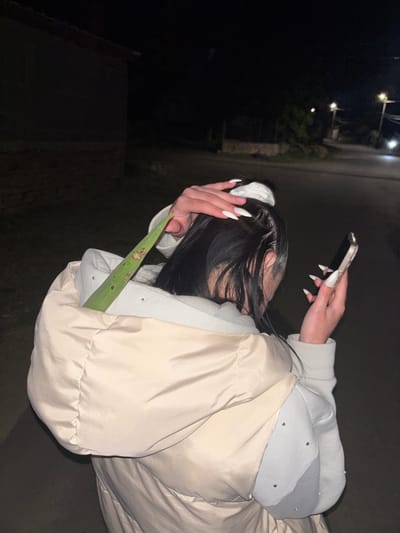 A person with long, dark hair is pictured from the side, facing away from the viewer. They are holding a smartphone in their right hand and have their left hand near their head. The person is wearing a light-colored puffer jacket and a light-colored hoodie with small embellishments. They are also holding a long, green plant leaf. The background is dark, suggesting it is nighttime on a street with streetlights visible in the distance. The exact location in Goritsa, Bulgaria, cannot be determined from the image.