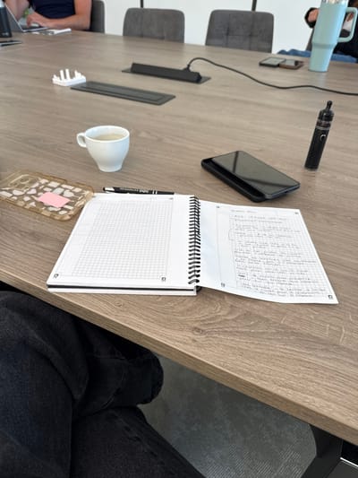 A spiral-bound notebook is open on a wooden table. The top page contains handwritten notes in French, and the bottom page is blank with a grid pattern. A black pen lies on the notebook. A black smartphone and a vape pen are on the table. A white coffee cup with coffee and a phone case are on the table as well. A chair is visible along the left side of the table, and a person's lap and legs are visible on the right side. The location is Les Arcs, France.