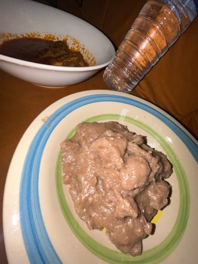 A meal is presented with a plate of brown, porridge-like food, possibly a starchy staple. A separate white bowl contains pieces of meat stewed in a dark red sauce. A plastic water bottle sits nearby. The plates rest on a surface with a warm, brown tone, likely a table or couch. The scene is brightly lit.