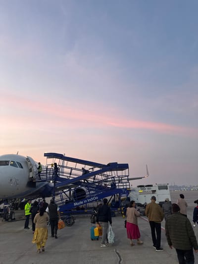 Passengers are boarding an IndiGo plane via a blue staircase. The plane's nose and a portion of the fuselage are visible. The sky shows a gradient of light blue and pink hues, suggesting early morning or late evening. Some airport vehicles and ground staff are near the plane. The scene appears to be at a busy airport terminal.
