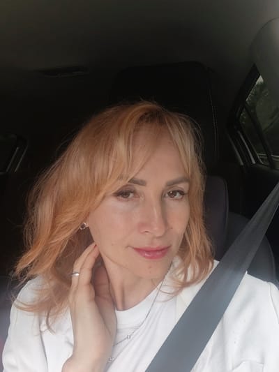 A woman is taking a selfie inside a car, likely in the city of Belka, Russia. She has blonde hair with reddish-blonde highlights and is wearing a white t-shirt. She is touching her neck with her left hand, revealing a ring on her finger and a necklace. A seatbelt is visible diagonally across her chest, secured over her t-shirt. The interior of the car appears dark, with hints of the outside environment visible through the windows, suggesting it is daytime.