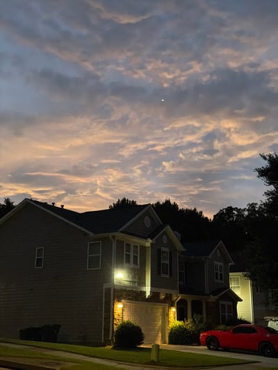 The image depicts a twilight scene in Buford, United States, featuring a residential street with townhouses. The sky is a canvas of muted blues and oranges, with scattered clouds. In the foreground, a row of two-story townhouses with neutral-toned siding and dark roofs are visible. One townhouse has a prominent garage with a lit interior, and a red sports car is parked in the driveway. Landscaping includes green lawns and bushes. The overall atmosphere is calm and residential.