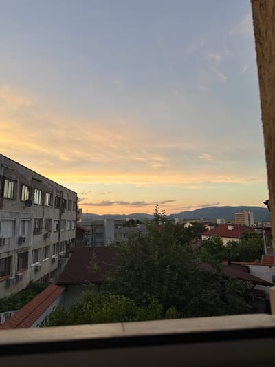 The image captures a sunset over the city of Kyustendil, Bulgaria. The sky is painted with soft hues of orange and yellow, transitioning to a pale blue towards the top, with wispy clouds scattered across. In the foreground, a portion of a textured, light-colored wall frames the view. To the left, a multi-story concrete building with numerous windows, some with air conditioning units, stands prominently. The building appears weathered, with peeling paint and visible plaster damage. Adjacent to this building, several houses with red-tiled roofs are visible, interspersed with lush green trees and foliage. In the background, other buildings of varying heights can be seen, including a taller, modern-looking structure. Beyond the city, rolling hills or mountains form a distant silhouette against the twilight sky. The scene is devoid of any people or visible activity, presenting a static, urban landscape at dusk.