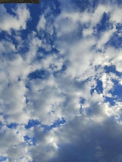 The image displays a sky observed over the city of Rosh HaAyin, Israel. The scene is dominated by a clear blue sky interspersed with various cloud formations. Numerous cumulus and stratocumulus clouds are visible, varying in size, shape, and density. Patches of bright white clouds, indicative of direct sunlight, are juxtaposed with areas of darker grey clouds, suggesting thickness or shadowing. Sunlight appears to be illuminating the cloud tops and edges from an unseen source. A small, partial architectural element, possibly a concrete or stone overhang or building edge, is visible at the very top of the frame, slightly left of center, indicating the upward perspective of the photograph. No discernible people, specific man-made objects within the sky, or active interactions are present. The environment depicted is exclusively the atmospheric sky.