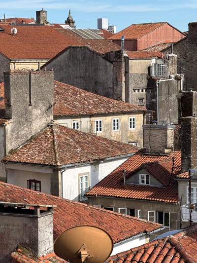 This is an aerial view of the rooftops of the historic city of Santiago de Compostela, Spain. The scene is dominated by terracotta tiled roofs, interspersed with stone and stucco buildings. Several chimneys, some made of brick and others of stone, punctuate the skyline. Modern elements like satellite dishes and antennas are visible, contrasting with the ancient architecture. The sky is clear and blue. No people are visible in the image. The overall impression is of a densely populated urban environment with a long history.