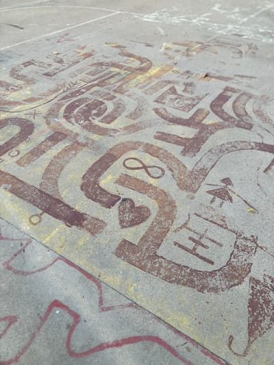 The image depicts a concrete surface, likely a playground or basketball court, adorned with colorful chalk drawings. The chalk art is rendered in shades of brown and yellow, with some red chalk lines also visible. The drawing appears to be a maze or abstract design, featuring curved and straight lines that form various shapes and patterns. Within the chalk art, there are small, stylized drawings, including a heart symbol and an infinity symbol. Further examination reveals a simplified depiction of a person holding what appears to be a broom or stick, and some letterforms that resemble "HI". The ground is textured and shows signs of wear, suggesting it is an outdoor public space. The lighting indicates it is daytime, with some shadows cast on the surface. The context provided indicates this scene is located in the city of Badalona, Spain.