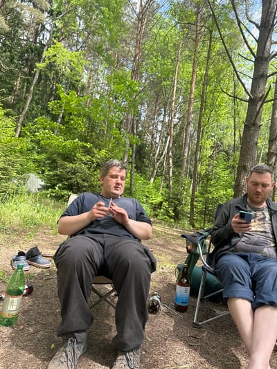 Two men are sitting in folding chairs in a wooded area. The man closer to the viewer is sitting on the right, wearing dark pants and a t-shirt. He's holding something small between his fingers. To his right, on the ground, are shoes, a bottle, and a partially visible can. The other man is seated behind and slightly above him, looking at a phone. He's wearing shorts and a t-shirt under a jacket. Next to him, on the ground, are bottles. The area is a forest with green foliage and tall trees. The location is Юзуфоўскі сельскі Савет, Belarus.
