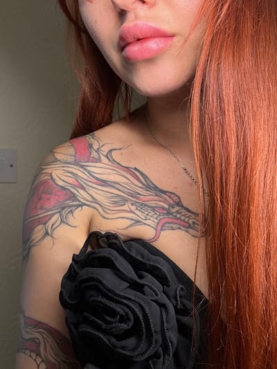 The image shows a close-up of a person with bright red hair. The person has pink lipstick on and a tattoo of a dragon on their shoulder and upper arm. They are wearing a black garment with a ruffled texture over their chest. The background is a plain, light-colored wall with a white electrical outlet. The lighting is soft, casting a slight shadow on the left side of the image. There is no discernible location in Oldham, United Kingdom, visible in the image.