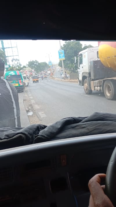 A view from inside a vehicle captures a bustling road scene. A large truck with a yellow cylindrical component is on the right. A bus and other vehicles are visible further down the road, with billboards lining the sides. The dashboard and the driver's hand on the steering wheel are in the foreground, creating a sense of being inside the vehicle.