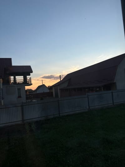 The image is a vertical, low-angle shot capturing parts of buildings and a grassy area under a clear, pale blue sky. The sky takes up a significant portion of the frame, with a hint of sunlight peeking through from the right side, casting a warm glow on some scattered clouds.
In the foreground, a dark, multi-story building with a reddish-brown, sloped roof dominates the left side. This building appears to have multiple levels and balconies, with some windows visible. Adjacent to it, to the right, is a lighter-colored building with a more complex structure, featuring balconies and open framework elements. Further back, another building with a prominent red roof is visible.
A fence runs horizontally across the middle ground, separating the buildings from a dark, densely-mowed grassy area on the right. The grass appears somewhat overgrown in places. The overall setting suggests a residential or industrial area, possibly in a rural or suburban environment.
The lighting suggests it is either early morning or late afternoon, given the warm tones in the sky. The weather appears clear and calm. No people are visible in the frame, and there is no visible text. The scene evokes a sense of quietness and stillness. The location context provided is Chemerne, Ukraine.