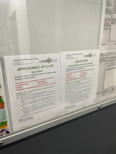 Two white printed notices, titled "APPEARANCE OF A LOT" and "CAR SPACES," are displayed in a transparent frame or case, affixed to a dark gray surface. The image is taken at an angle, with the text oriented sideways.
The "APPEARANCE OF A LOT" notice, on the left, outlines rules for balconies at Urban Edge Apartments in Kelvin Grove, Australia. It prohibits the storage of items other than typical outdoor furniture, specifically listing gym equipment, fridges, mattresses, clothes airers, and planter box greenhouses (above railing height) as forbidden. An extract from the body corporate by-laws details requirements for architectural code compliance, prohibits changing external appearance, hanging washing visible from outside, displaying signs, and placing non-balcony furniture on balconies. Residents are asked to remove such items.
The "CAR SPACES" notice, on the right, prohibits the storage of items other than vehicles (cars, motorbikes, trailers) in parking spaces. Specifically disallowed items include toolboxes, suitcases, furniture/beds, fuel cans, children's toys, mattresses, and unregistered/unroadworthy vehicles. Partial text from the body corporate by-laws regarding car parking is also visible.
Both notices provide contact details for "URBAN EDGE APARTMENTS KELVIN GROVE, 63 Blamey Street, Kelvin Grove Qld 4059, Building Management: (07) 3129 6409," and are issued by the "URBAN EDGE APARTMENTS Body Corporate Committee."