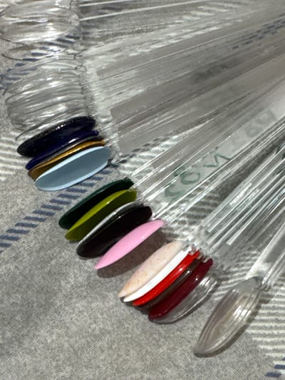 The image shows a set of clear plastic nail polish swatch sticks arranged on a grey textured surface. The swatch sticks are fanned out and each displays a different color of nail polish. From top to bottom and left to right, the visible colors include dark blue, gold, light blue, brown, dark green, olive green, black, pink, a speckled light color, white, red, and dark red. The lighting is overhead and creates highlights on the glossy nail polish colors. The background surface appears to be a fabric, possibly a blanket or textile. The scene is indoors. There is no indication of the city of Яромінскі сельскі Савет, Belarus, or any specific location within it.