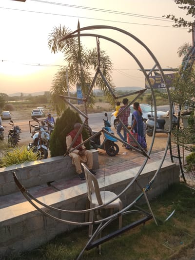 The image depicts a scene in Kondamaripalle, India. The foreground features a large, metal sculpture combining a crescent moon and a star. An adult male, dressed in traditional Indian attire (khaki pants, red overshirt), is seated on a concrete ledge next to the sculpture. Adjacent to him is a white plastic chair.

In the background, there is a road with parked motorcycles and cars. Two adults, one carrying a child, are walking beside a white car. The environment is outdoors, with a clear sky and a landscape of distant hills visible in the background. A building with signs is to the right of the frame. The time of day appears to be late afternoon or early evening, based on the lighting.