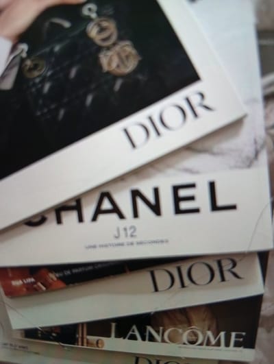 The image shows a stack of fashion magazines. The top magazine features the brand name "DIOR" prominently displayed. Below it, another magazine showcases "CHANEL" along with "J12 UNE HISTOIRE DE SECONDES". A third magazine displays "DIOR" again, and the bottom magazine visible has "LANCOME" printed on it. There are also some partial product names visible like "EAU DE PARFUM ORIG" and "DIUA LIPA". The magazines are layered on top of each other, creating a slight angled perspective. The lighting appears to be indoor, with no specific time of day or weather cues discernible. The focus is on the branding and text of the magazines, suggesting a commercial or retail setting, possibly a display or a stack of personal items. No people are clearly visible, only a hint of a hand and arm on the top magazine.