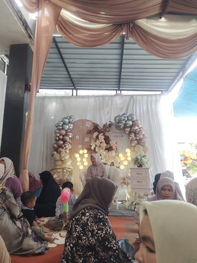 This image captures a celebratory event, likely a baby's full month celebration or a similar family gathering, taking place indoors with an open-air roof structure. The scene is decorated with a backdrop featuring a white curtain, an arched wooden frame adorned with floral arrangements and balloons in shades of silver, champagne, and cream. A string of fairy lights enhances the festive atmosphere.
Several people are present, including adults and children, mostly dressed in casual attire with many wearing hijabs. In the center of the frame, a woman in a light purple dress and hijab is seated on a white couch, looking towards the camera and smiling. Children are scattered around the area, with one young boy in the foreground holding a pink and green toy. Another child is seated on the floor, looking attentively at the scene.
The decor suggests a happy occasion, with the balloons and flowers creating a welcoming and joyful ambiance. The lighting indicates it is daytime, with natural light filtering in from the sides. The setting appears to be a temporary event space, possibly a community hall or an outdoor pavilion, common in Indonesian celebrations. The presence of balloons and a decorated backdrop points to a planned event rather than a casual get-together. There is no visible text on the balloons, but the archway has text which appears to read "Gheeziya zhafirah." The overall mood is one of festivity and familial warmth.