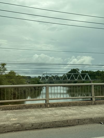 An outdoor, eye-level shot captures a cloudy day in Nam Đông Hà Ward, Vietnam. The foreground features a sidewalk made of grey square tiles with a textured pattern, bordered by a concrete curb and a grey metal railing. Beyond the railing, a calm river reflects the overcast sky and the lush green trees lining its banks. A metal truss bridge, its triangular structure prominent, spans the river in the mid-ground. Several dark electrical wires are strung horizontally across the top portion of the frame, obscuring parts of the sky and the distant landscape. The overall atmosphere is subdued, with muted colors and soft lighting characteristic of a cloudy day.