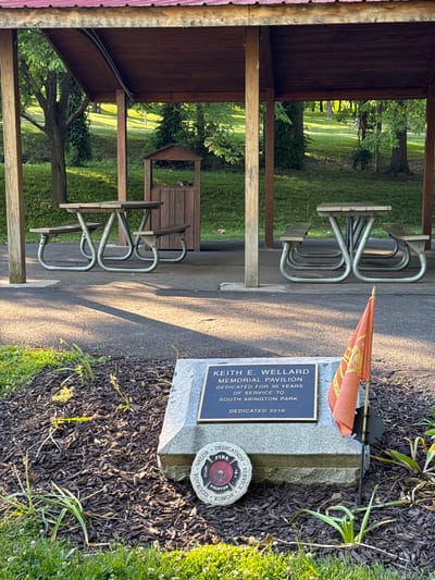 The image shows a memorial pavilion and picnic tables in a park setting, likely South Abington Park in South Abington Township, United States of America. In the foreground, a stone monument with a bronze plaque reads "KEITH E. WELLARD MEMORIAL PAVILION DEDICATED FOR 30 YEARS OF SERVICE TO SOUTH ABINGTON PARK DEDICATED 2019". A small orange flag with a crest is planted in front of the monument. To the left of the monument is a circular stone emblem with the words "VALOR COURAGE HONOR SERVICE FIRE FIGHTER DEDICATION". Behind the monument, several picnic tables with benches are visible under a wooden pavilion with a reddish-brown roof. The ground is paved with asphalt and covered with mulch in the memorial area. Trees and grassy slopes form the background, with sunlight filtering through the leaves.